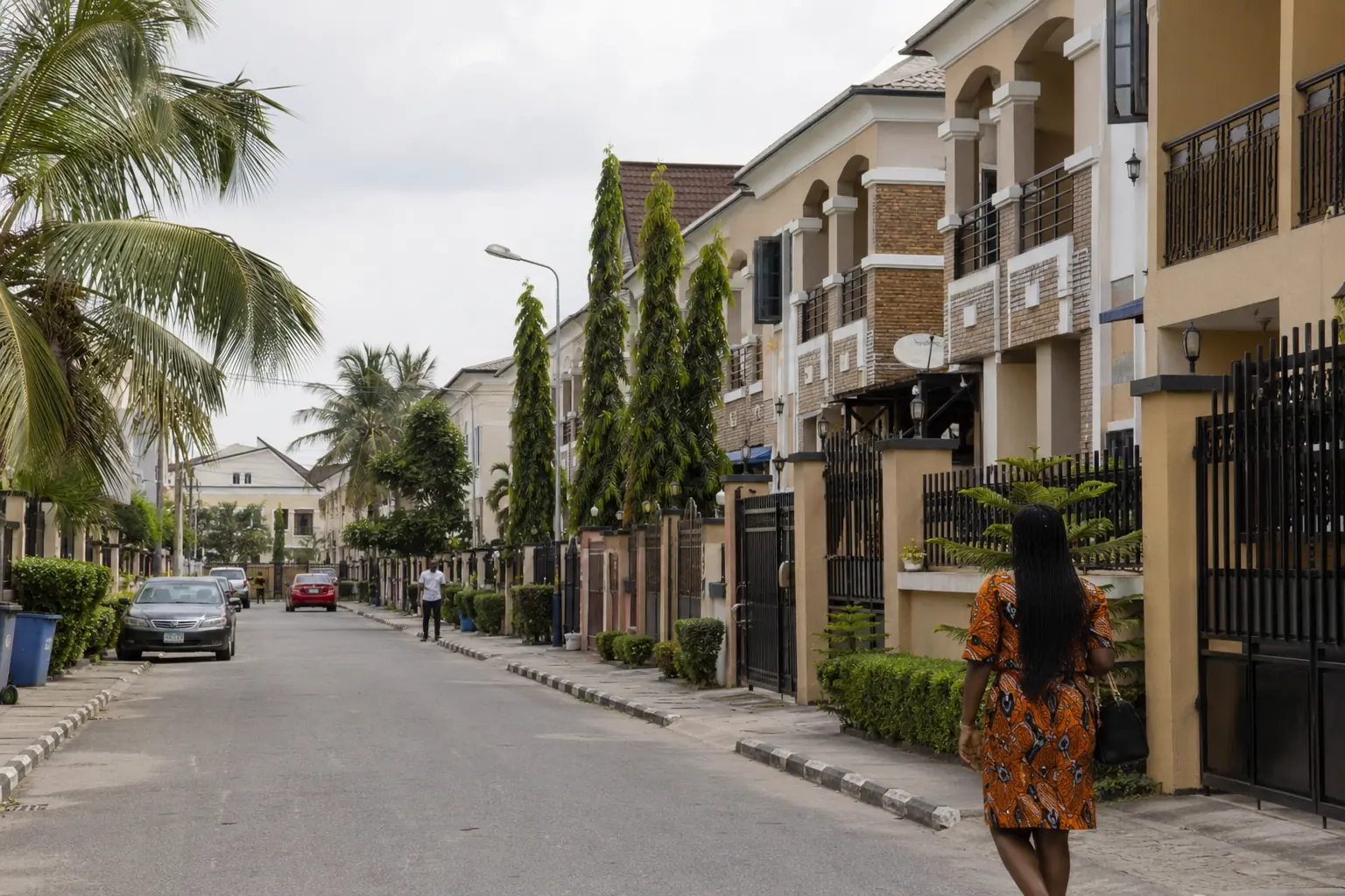 A quiet residential street in a Nigerian estate, lined with gated two-storey homes and palm trees.